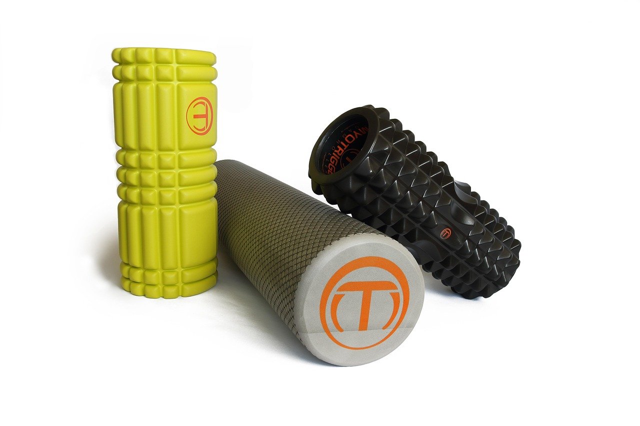 rolo de espuma (foam roller) texturizado apoiado verticalmente ao lado de um tapete de exercício dobrado, tons neutros, luz suave